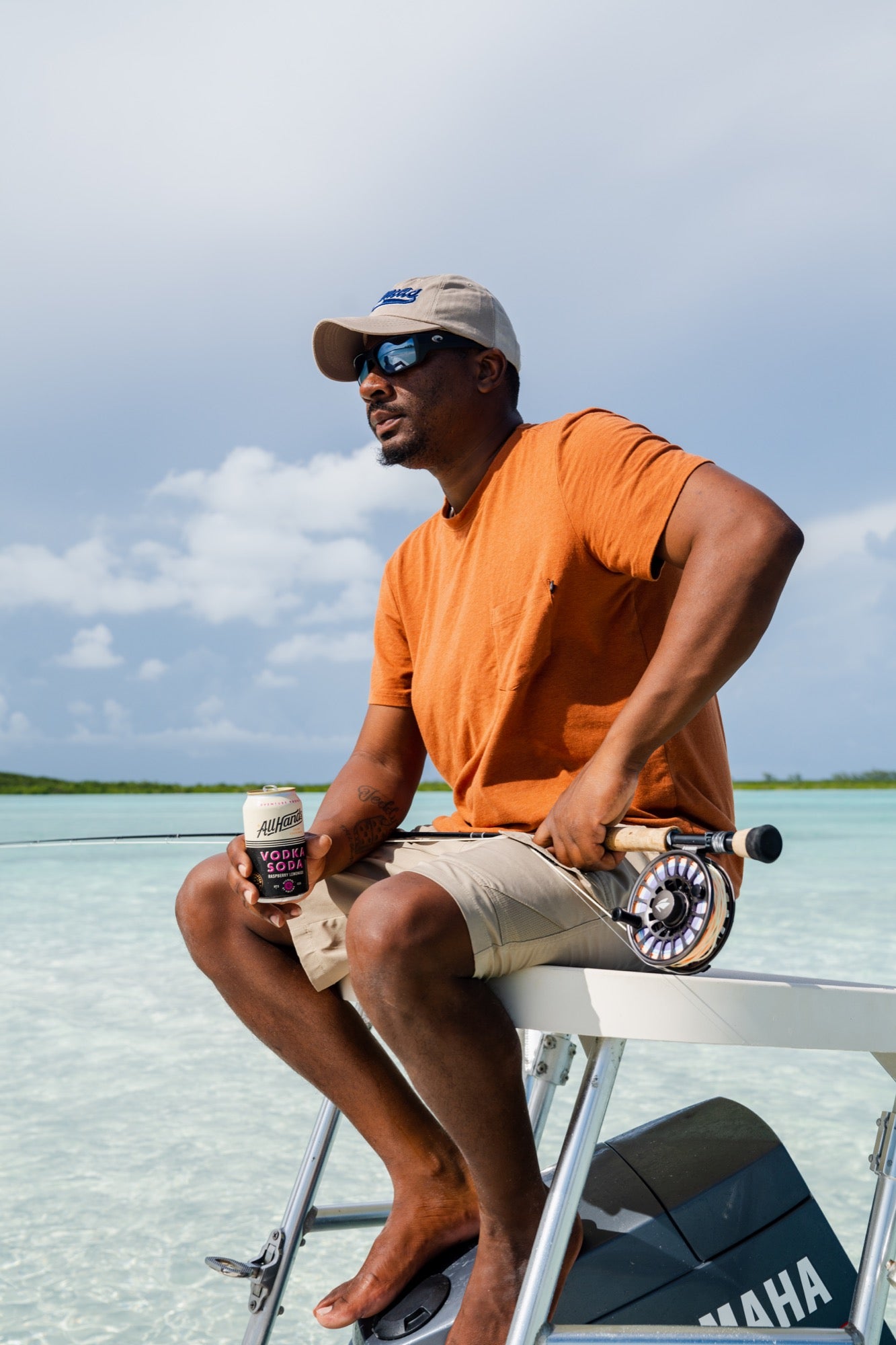 A man in a Choona Panhandle Pocket Tee, khaki shorts, and a beige cap sits on a boats edge in shallow, clear water, holding a fishing reel and coffee cup under a cloudy sky.