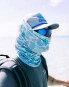 Wearing a Choona Grayton Sun Mask in light blue pattern, blue mirrored sunglasses, and a light gray cap, a person with a backpack faces the ocean outdoors on a sunny day.