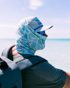 Wearing a Choona Grayton Sun Mask, hat, and sunglasses, a person gazes over clear shallow ocean water beneath a blue sky. A backpack strap on their shoulder signals readiness for outdoor adventure and sun protection.