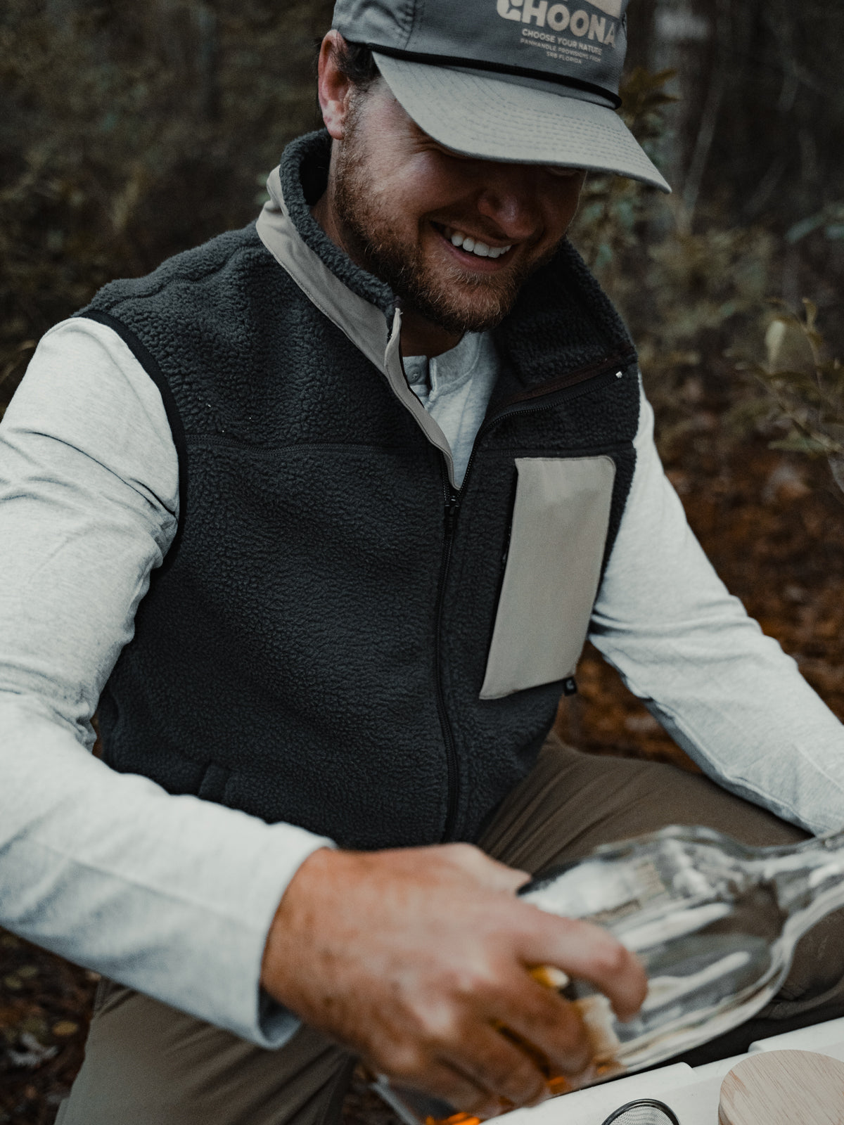 A man in a gray cap and Choona Pt. Washington Sherpa Vest smiles while pouring a drink outdoors amid greenery. The high-pile fleece of the vest delivers cozy warmth for all your adventures.