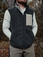 A person in a Choona Pt. Washington Sherpa Vest with a contrasting chest pocket, light long-sleeve shirt, and khaki pants leans against a tree outdoors, hands in pockets.