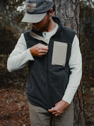 A man in a Choona Pt. Washington Sherpa Vest with Durable Water Repellent finish, a light long-sleeve shirt, and a cap stands by a tree, zipping his vests chest pocket as he looks down, his face partially hidden.