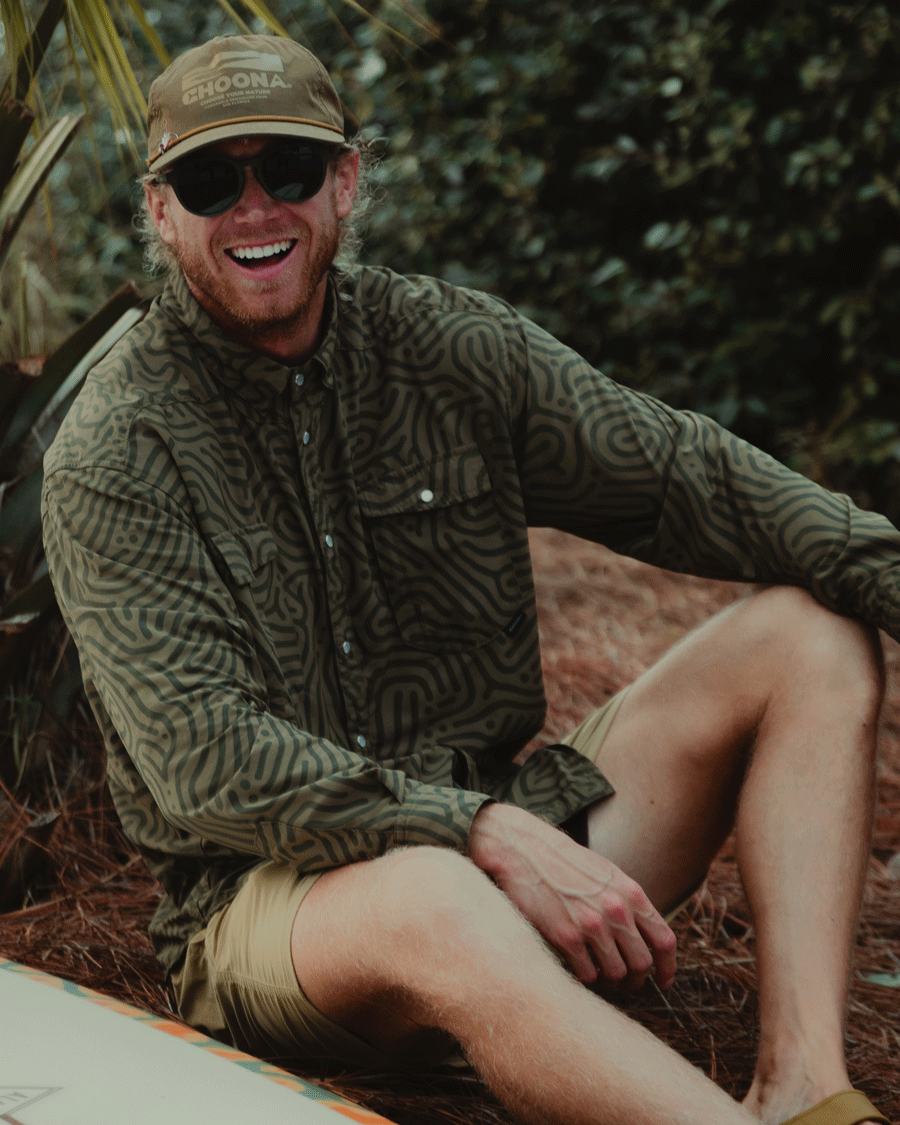 A smiling man sports sunglasses, a cap, and the Choona Panhandle Pearl Snap shirt as he relaxes outdoors among palm leaves and bushes, capturing the laid-back spirit of the Florida Panhandle.