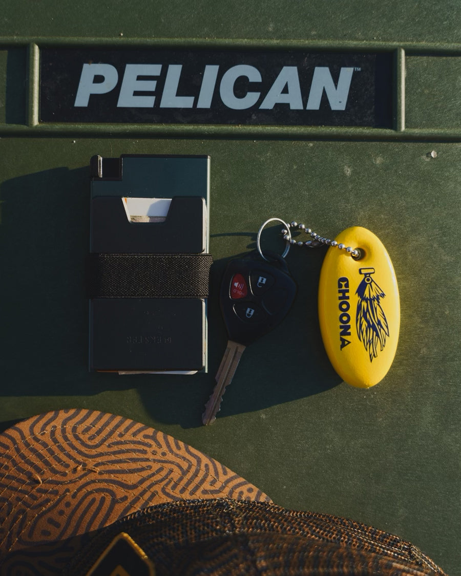 A wallet, car keys with a yellow Choona Floating Keychain by Choona, and part of a patterned hat rest in style on a green cooler.