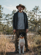A man in outdoor clothes and a Choona Skipper Shacket stands in a grassy field holding a leash, with his black and white dog sitting in front. Pine trees and blue sky suggest crisp, cold weather in the background.