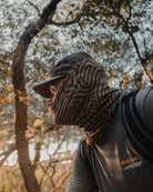 Wearing the Choona Grayton Sun Mask, a cap, and a backpack, a person in a CHOONA shirt stands in a sunlit forest. Sunlight filters through the trees, highlighting outdoor protection.