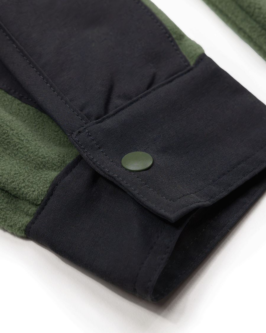 Close-up of the Choona Skipper Shacket sleeve in dark green and black recycled polyester, featuring a green snap button on the cuff that highlights the contrasting fabric textures.