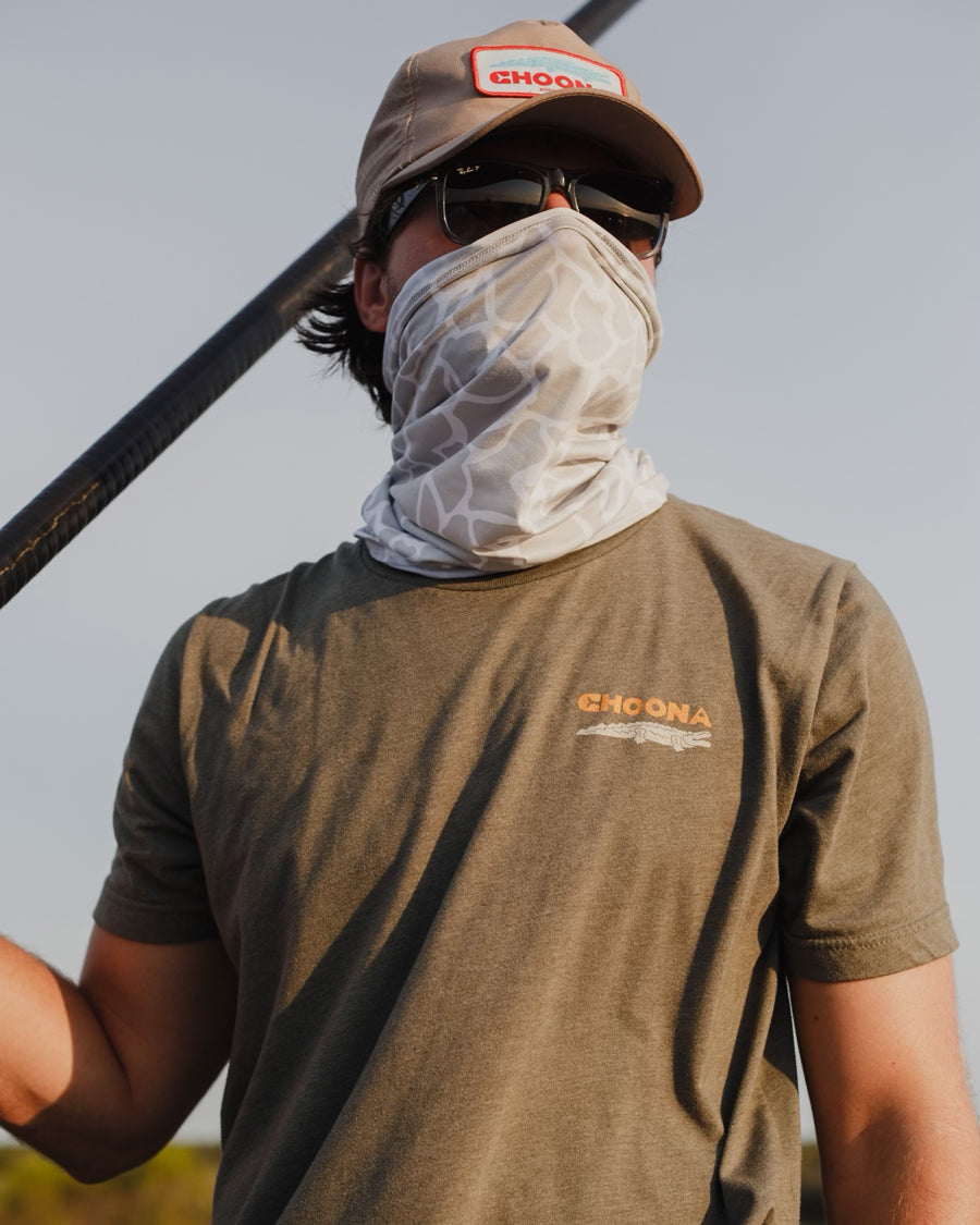 Outdoors under a clear sky, someone in sunglasses, a beige cap, an olive green Choona t-shirt, and the ultra-lightweight Choona Grayton Sun Mask stands holding a pole, demonstrating effective outdoor protection.