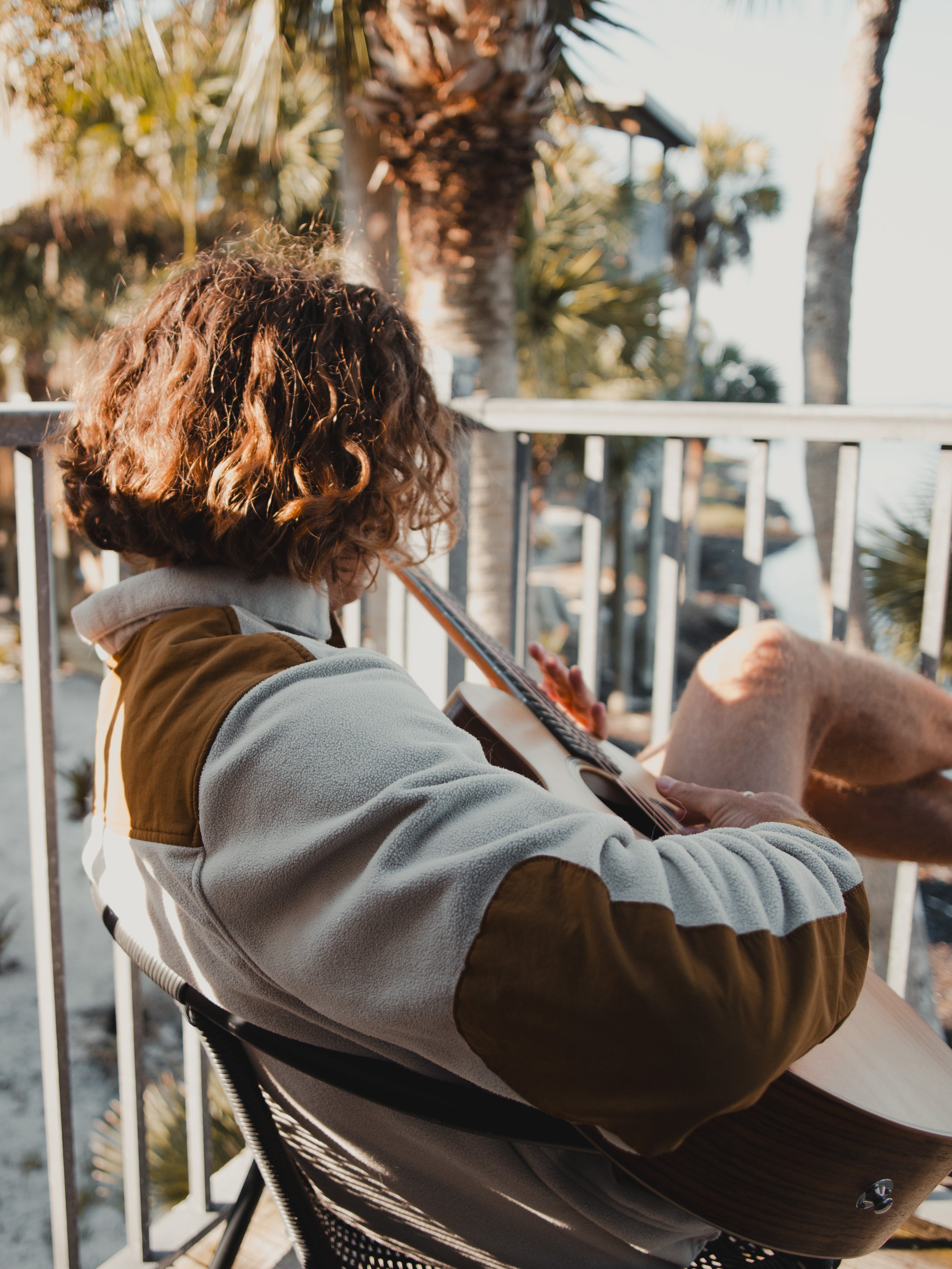 A person with curly hair sits outdoors under palm trees, playing an acoustic guitar and wearing the Choona Skipper Shacket, made from recycled polyester for a relaxed, warm vibe.