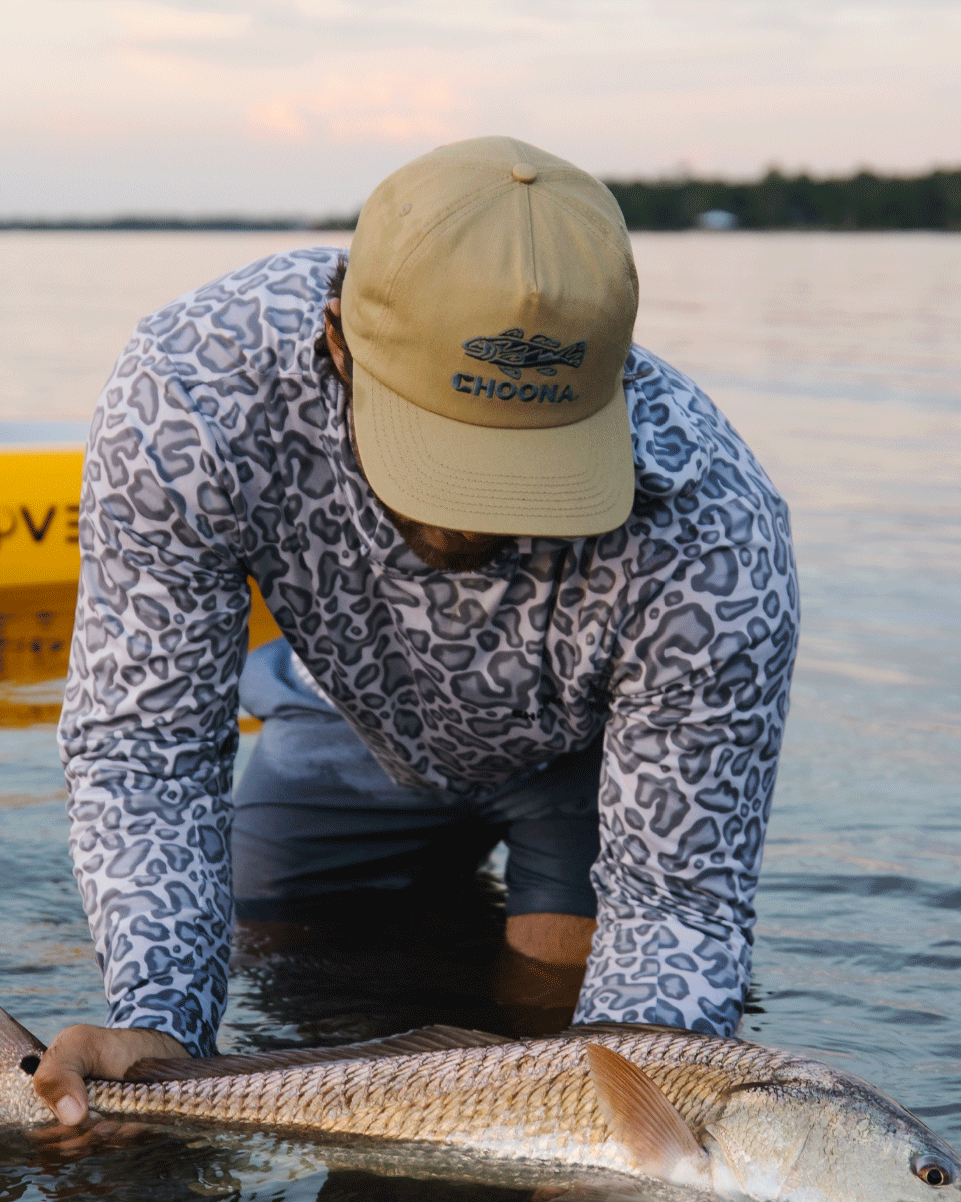 Wearing a Choona Redfish Snapback, a person in a patterned long-sleeve shirt leans over shallow water holding a big fish, with calm water and part of a yellow kayak visible in the background.