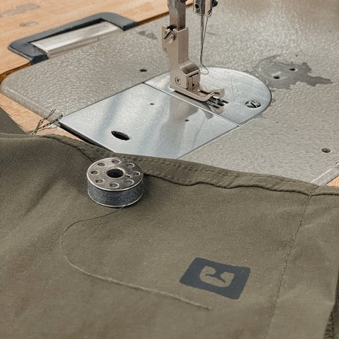 A close-up of a sewing machine with olive green fabric under the needle. A metal bobbin with thread rests on the fabric, which features a small black patch with a minimalist logo on it.
