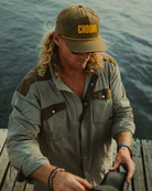A person with long blond hair wears a Choona brown cap with yellow text and the Panhandle Pearl Snap shirt, standing on a wooden dock in the Florida Panhandle, looking down while holding an object.