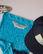 A blue ultra-breathable Choona Grayton Sun Mask with a fish illustration sits beside a black cap featuring a colorful patch, all arranged on a light surface with a metal hinge—ideal for protection in extreme conditions.