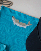 A close-up of the Choona Grayton Sun Mask, featuring ultra-breathable blue fabric with a wavy pattern and a light gray fish graphic with a white belly, shown next to a dark blue cap. A metallic fixture appears at the top left.