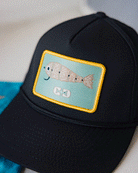 A close-up of the Choona X Dent Redfish Snapback by Choona, a black cotton twill cap with a rectangular patch showing an embroidered smiling fish and two fishhooks, all outlined in yellow.