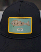 The Choona X Dent Redfish Snapback by Choona is a black cotton twill cap with a rectangular patch showing a cartoon fish with scales and a hook-shaped mouth on light blue, bordered in yellow.