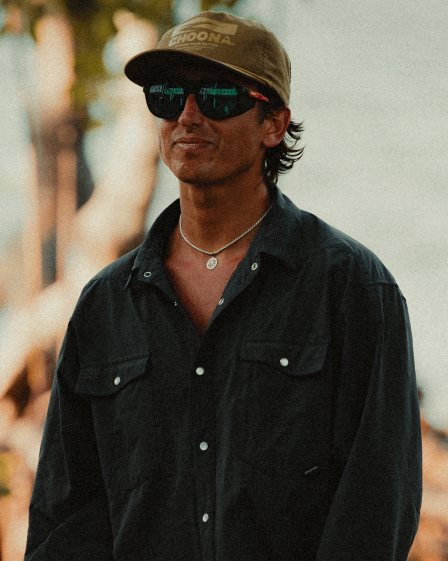 A person in a brown cap, dark sunglasses, and a black Choona Panhandle Pearl Snap smiles outdoors. The blurred background of trees and people hints at a relaxed Florida Panhandle coastal cowboy vibe.