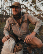 A man in a Choona Panhandle Pearl Snap shirt, hat, and neck gaiter smiles while crouching outdoors with camping gear among tall pines—capturing the spirit of a true coastal cowboy in Florida’s Panhandle.