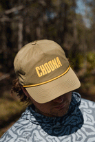 On a coastal adventure, someone sports the Choona Wavy Snapback—a khaki quick-drying cap with bold yellow CHOONA lettering—paired with a patterned gray hoodie as the brim shades their face.