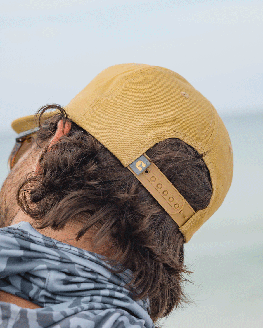 A person wearing a Choona Redfish Snapback—a yellow cotton canvas cap—paired with a patterned scarf faces a blurred outdoor background. The photo, taken from behind, highlights the cap and the persons wavy hair.