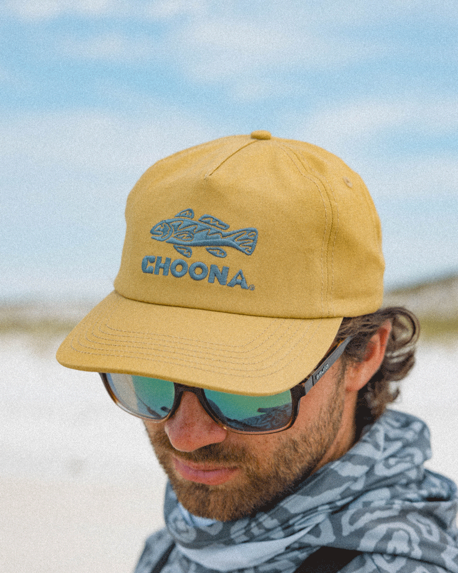 A man stands outdoors against a bright, sandy background wearing the Choona Redfish Snapback cap, featuring the redfish logo and “CHOONA” embroidery. He pairs it with reflective sunglasses and a patterned neck gaiter.