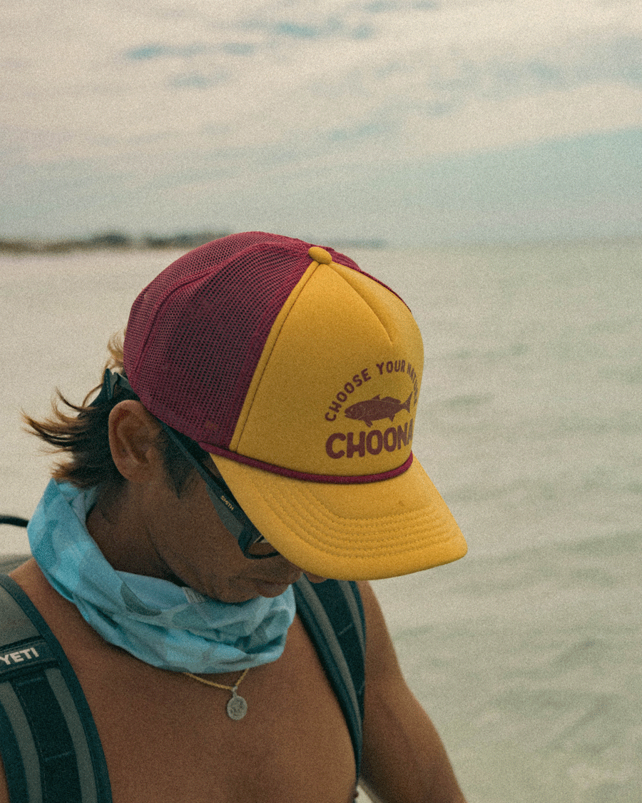 A person in sunglasses, a blue neck gaiter, and a Choona Redfish Foam Snapback stands by the water, looking down.