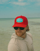 A man with light brown hair and a beard wears the Choona Breezy Snapback - Red, sunglasses, and a light gray hoodie while standing on a beach with shallow water beneath a cloudy sky.