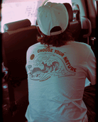 A person with long hair and a white cap sits in a vehicle, seen from behind. They're wearing the Choona Armachillo Graphic Tee, featuring waves, a sun, and "CHOOSE YOUR NATURE" on the back—crafted for comfort and style.