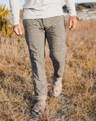 Wearing Choona’s Drifter Pant with an abstract pattern and four-way stretch, a person strolls through dry grass outdoors as sunlight creates shadows on the ground.