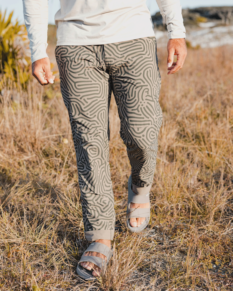 Wearing Choona’s Drifter Pant with an abstract pattern and four-way stretch, a person strolls through dry grass outdoors as sunlight creates shadows on the ground.