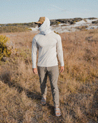 A person stands in a grassy field wearing a light long-sleeve shirt, Choona Drifter Pant with four-way stretch, sandals, a cap, sunglasses, and a white cloth wrapped around their head and neck for sun protection.