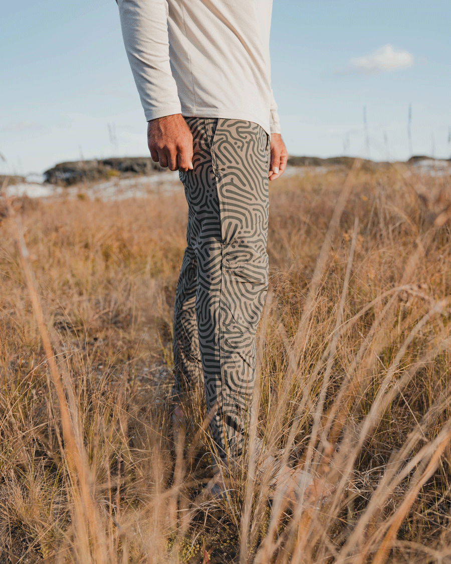 A person stands in tall, dry grass wearing a white long-sleeve shirt and Choonas Drifter Pant, which has a maze-like pattern and four-way stretch. Behind them are rocks and a blue sky with scattered clouds.
