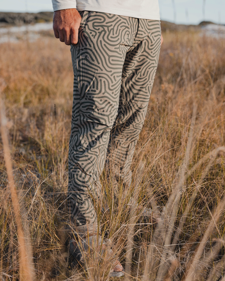 A person stands in tall, dry grass wearing Choona’s Drifter Pant—beige with a bold wavy black abstract pattern and four-way stretch—paired with a long-sleeve white shirt; only the lower body is shown.