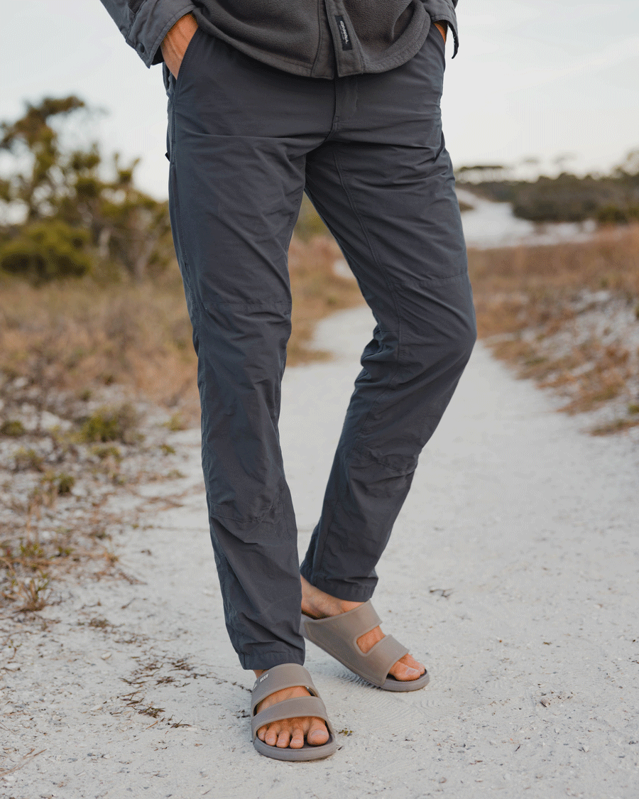 A person stands on a sandy path, wearing Choona Drifter Pant in dark gray with four-way stretch and gray slide sandals. Only their lower half is visible, set against sparse vegetation and a blurred sky.
