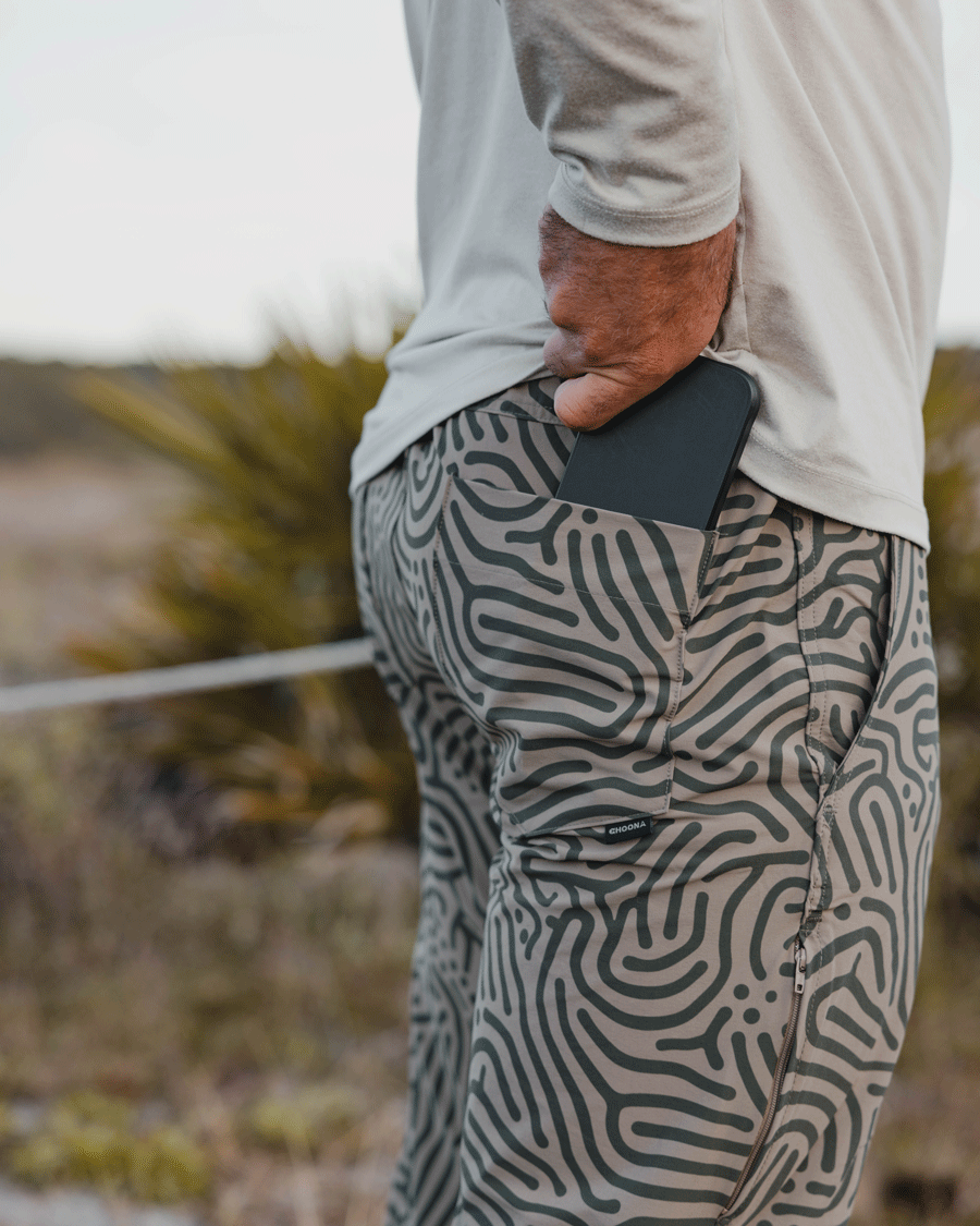 A person in Choona Drifter Pant with four-way stretch and a light long-sleeve shirt stands outdoors, slipping a black smartphone into their back pocket. Greenery appears in the blurred background.