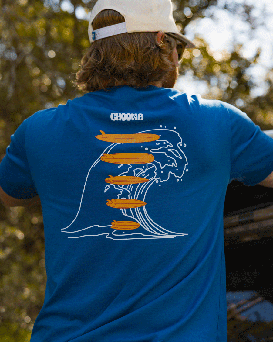 A person outdoors wears Choona's Swell Dweller Graphic Tee featuring a white wave outline and four orange surfboards under "CHOONA," paired with long hair and a white cap.