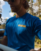 A man wearing sunglasses and a blue Swell Dweller Graphic Tee by Choona stands outdoors with trees and a cloudy sky in the background.