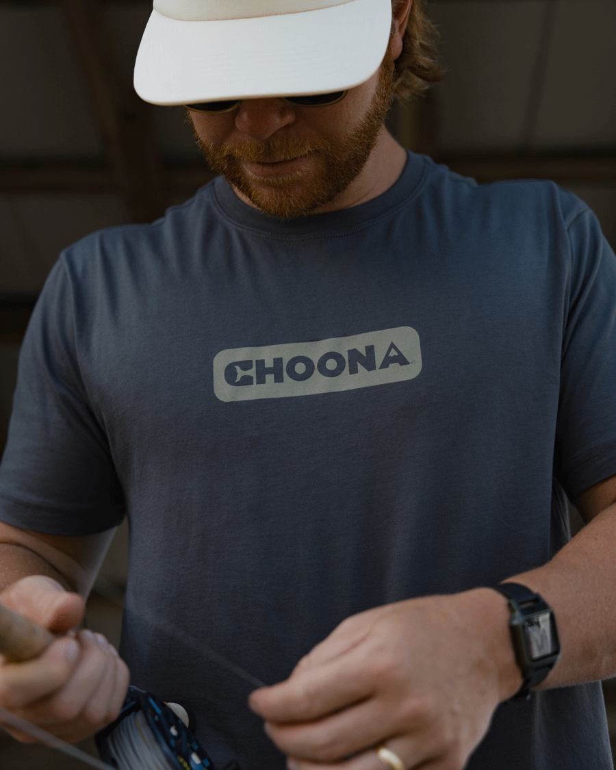 A bearded person in a blue Choona Logo Graphic Tee by Choona and a white cap looks down, holding a fishing rod and line with a watch on their left wrist—embracing the coastal lifestyle.