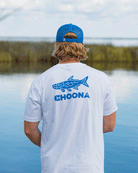 A person with blond hair, wearing a blue cap and a white Choona Tarpon Graphic Tee, stands by calm water and marsh grasses, facing away from the camera.