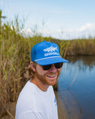 A smiling man with a beard and sunglasses wears a Choona Silver King Snapback and a white shirt, standing outdoors near tall grasses and water under a blue sky.
