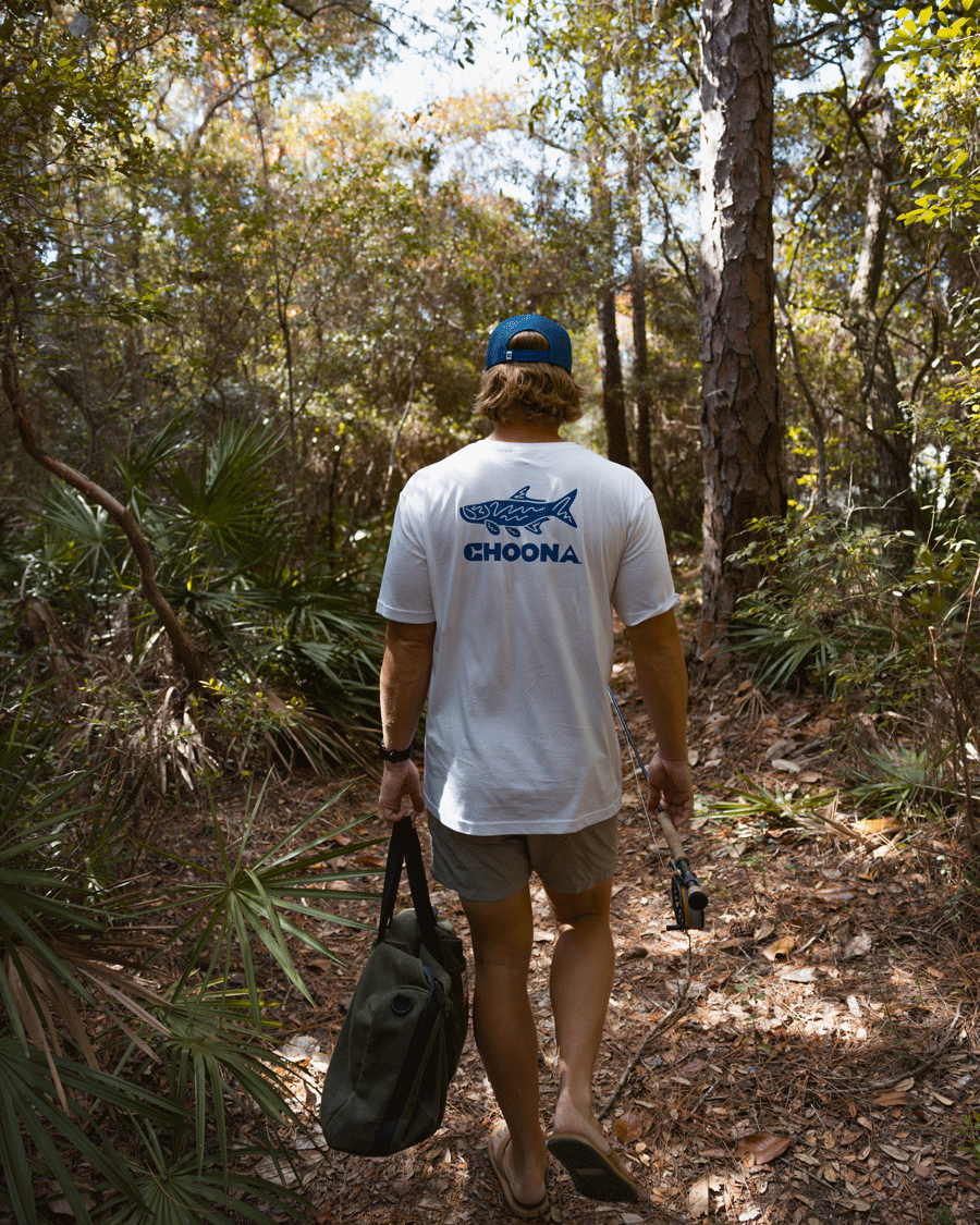 Wearing a Choona Tarpon Graphic Tee, shorts, and a blue cap, a person walks barefoot along a sunlit forest trail with a green bag and fishing rod, surrounded by trees and lush foliage.