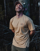 A man in a beige Choona SRB Natives Graphic Tee and cap leans against a wooden pole outdoors, eyes closed and smiling. Trees in low light create a peaceful Santa Rosa Beach evening atmosphere.