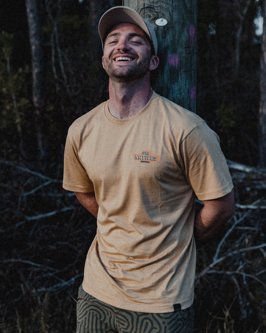 A man in a beige Choona SRB Natives Graphic Tee and cap leans against a wooden pole outdoors, eyes closed and smiling. Trees in low light create a peaceful Santa Rosa Beach evening atmosphere.