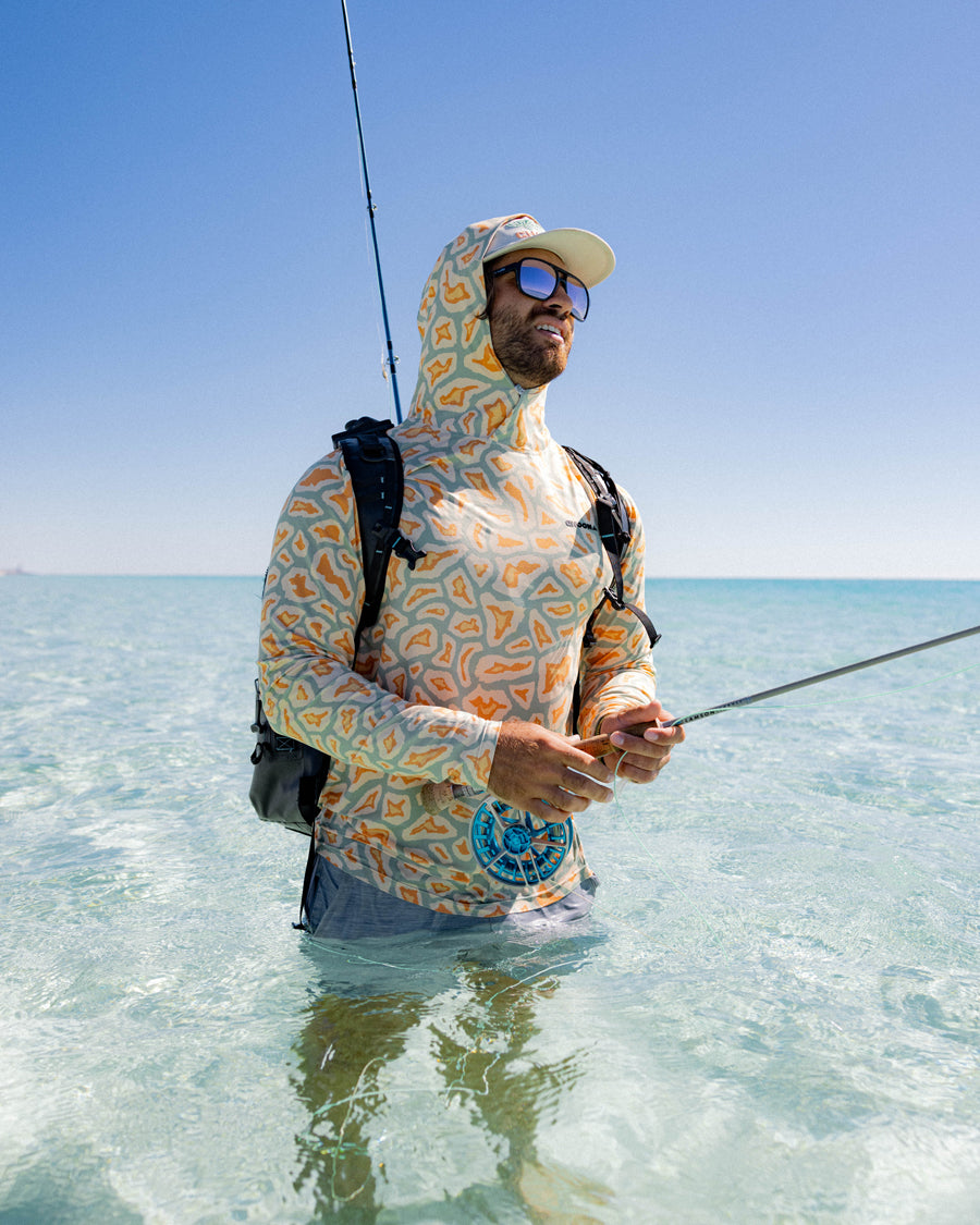 A man stands waist-deep in clear, shallow ocean water fishing with a rod. He wears the Choona Grayton Ultra-Lightweight Hoodie, sunglasses, a cap, and carries a backpack under a clear blue sky.