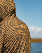 A person wears the Choona Grayton Ultra-Lightweight Hoodie, displaying its brown and black abstract, maze-like pattern while standing outdoors near water under a blue sky. Only the upper back and side of the hoodie are visible.