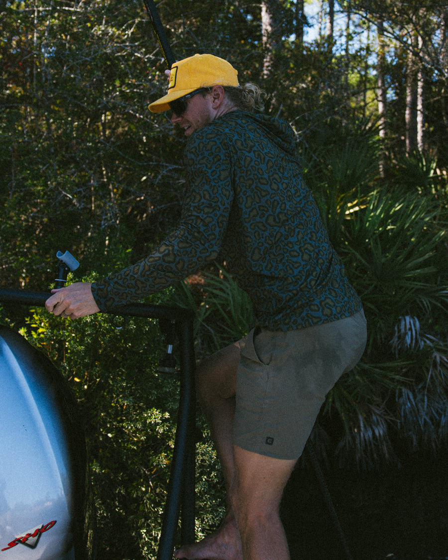 Wearing the Choona Grayton Ultra-Lightweight Hoodie and shorts, a person in a yellow cap stands outdoors holding a metal structure amidst trees and greenery.
