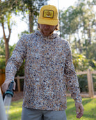 A man in a yellow cap and the Choona Grayton Ultra-Lightweight Hoodie waters plants with a garden hose outdoors, surrounded by trees and sunlight.