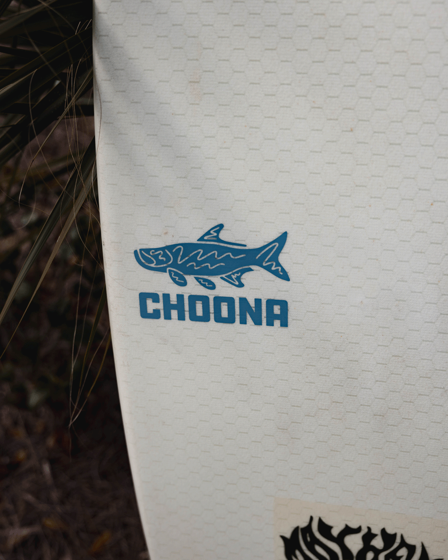A close-up of the Choona Tarpon Decal from Choona shows a blue tarpon graphic above CHOONA in blue on a white surfboard with a hexagonal pattern. Grass and foliage appear in the background.
