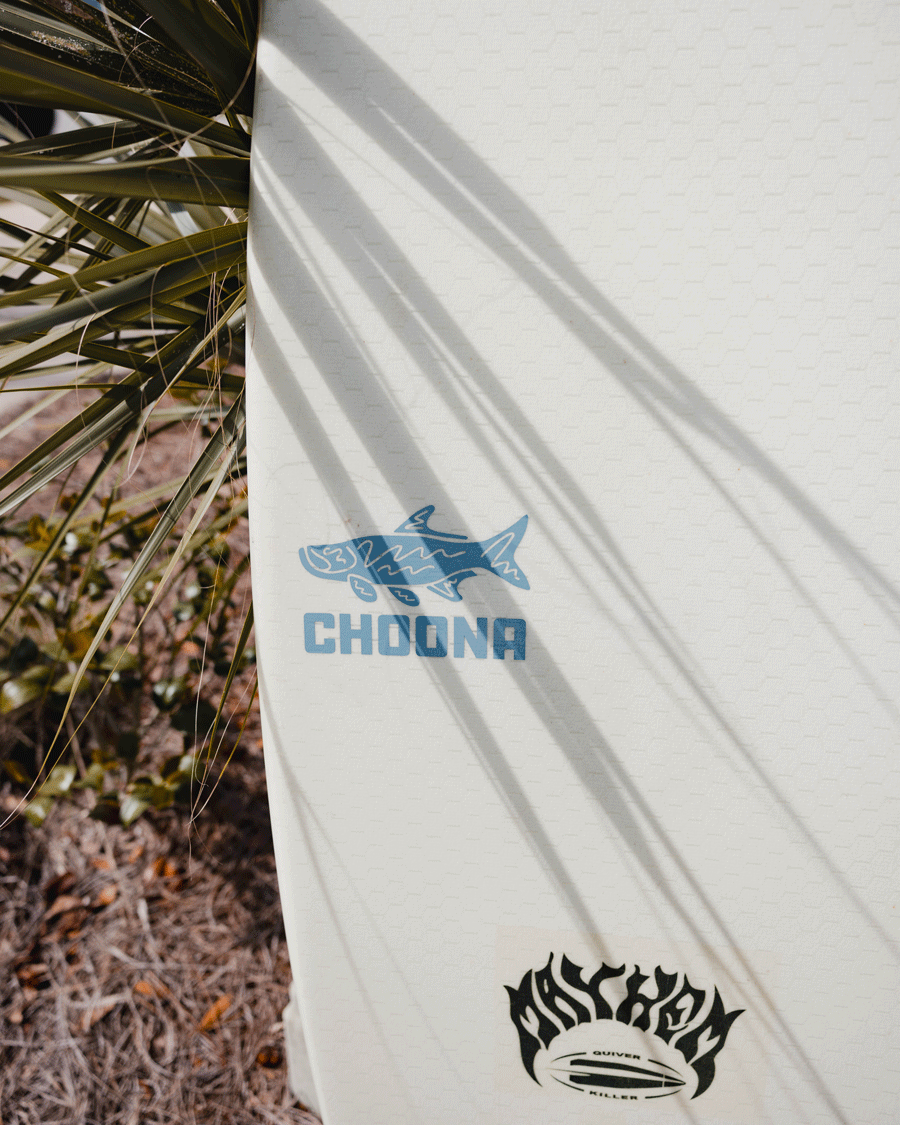 A close-up of a white surfboard features the Choona Tarpon Decal from Choona and a black MAYHEM logo. Palm leaves cast long shadows across the board, which sits among grass and plants in the background.