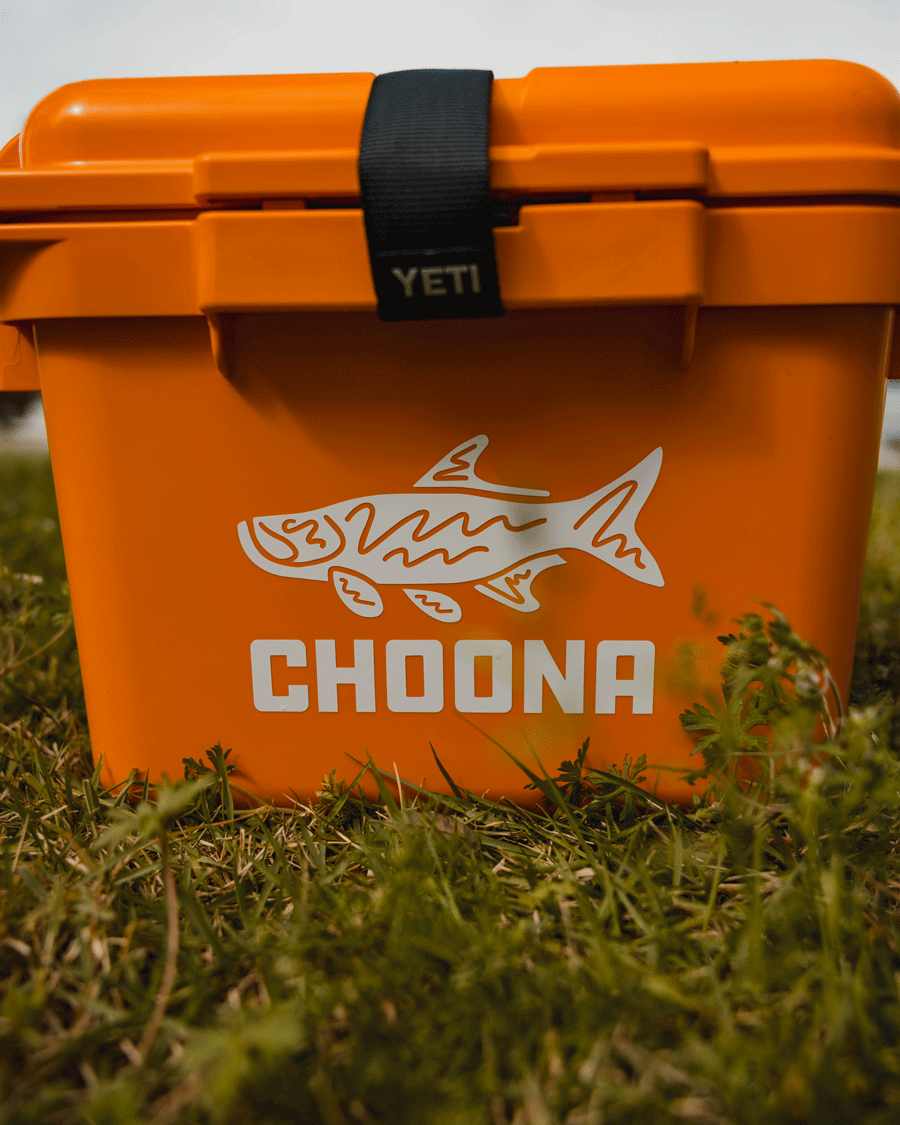 A Choona Tarpon Decal by Choona is displayed on an orange YETI cooler featuring a fish illustration and the word CHOONA, sitting on grass with green plants in the foreground.